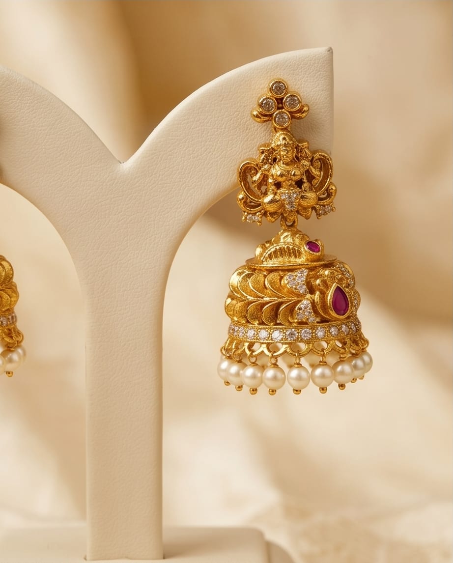 Gold jhumka earrings with pearls and a red gemstone on a beige stand.