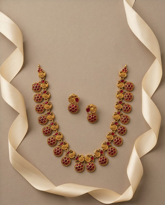 Gold necklace with red stones and matching earrings on a beige background with ribbons.