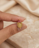 Gold pendant with pink gemstones held between fingers against a beige fabric background