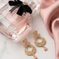 Gold earrings with pink stones next to a perfume bottle with a black ribbon on a light pink fabric background