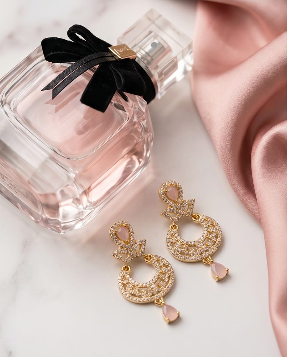 Gold earrings with pink stones next to a perfume bottle with a black ribbon on a light pink fabric background