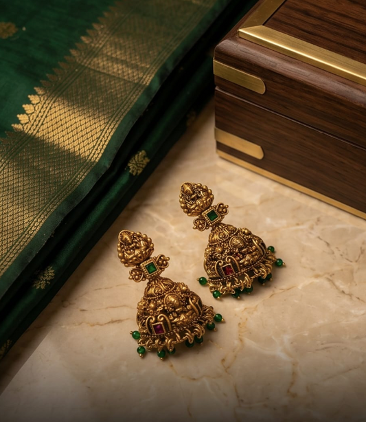 Gold earrings with green and red stones on a marble surface next to a saree and wooden box.