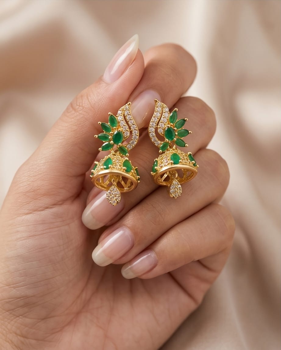 Gold earrings with green gemstones held in a hand against a beige background